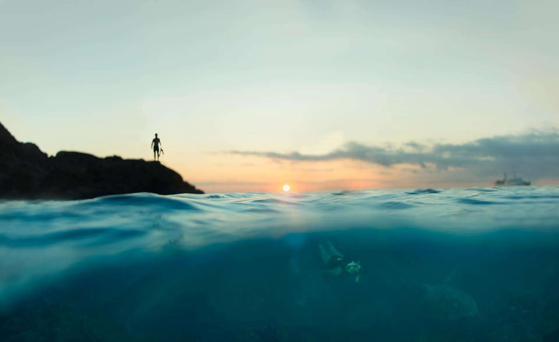 Sunset over the sea in the Galápagos, seen half above and half below the waterline, with a snorkeler and sea turtle beneath the surface